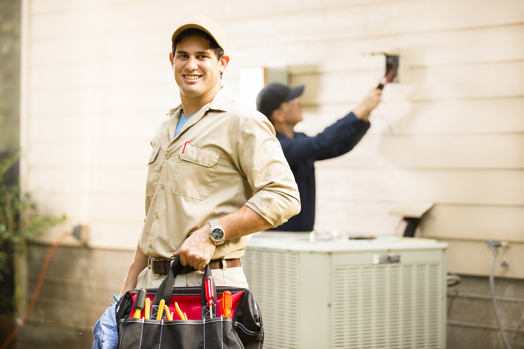 A person holding a tool belt with a person working on the siding of a house in the background.