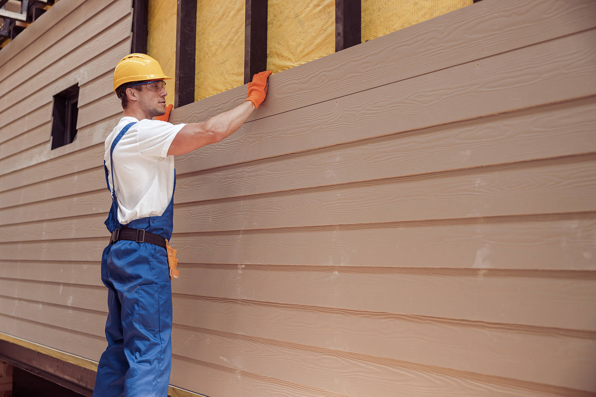 A person wearing a hard hat and blue overalls installing siding to a home.