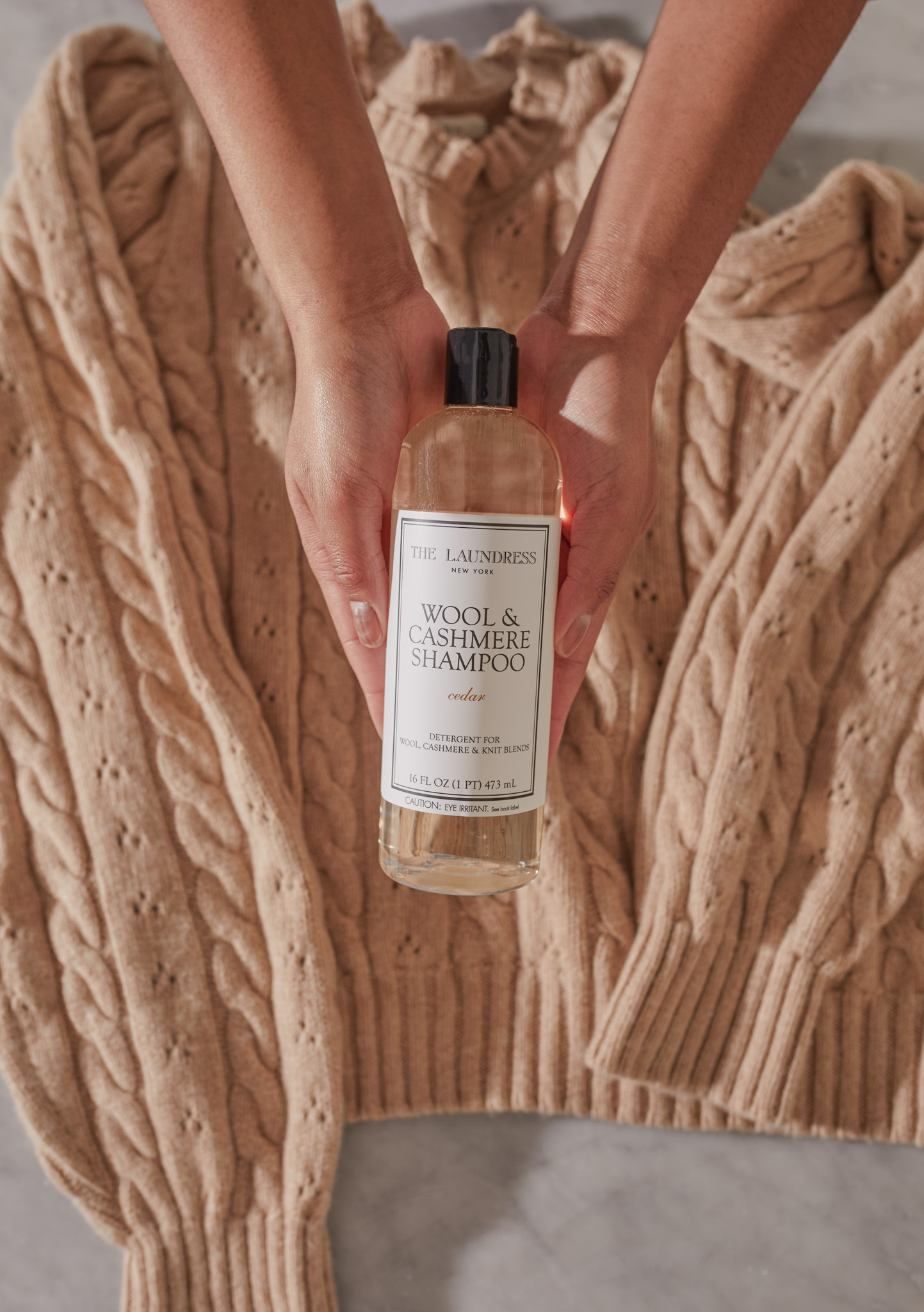 hands holding the Laundress Wool & Cashmere Shampoo over a tan cashmere sweater