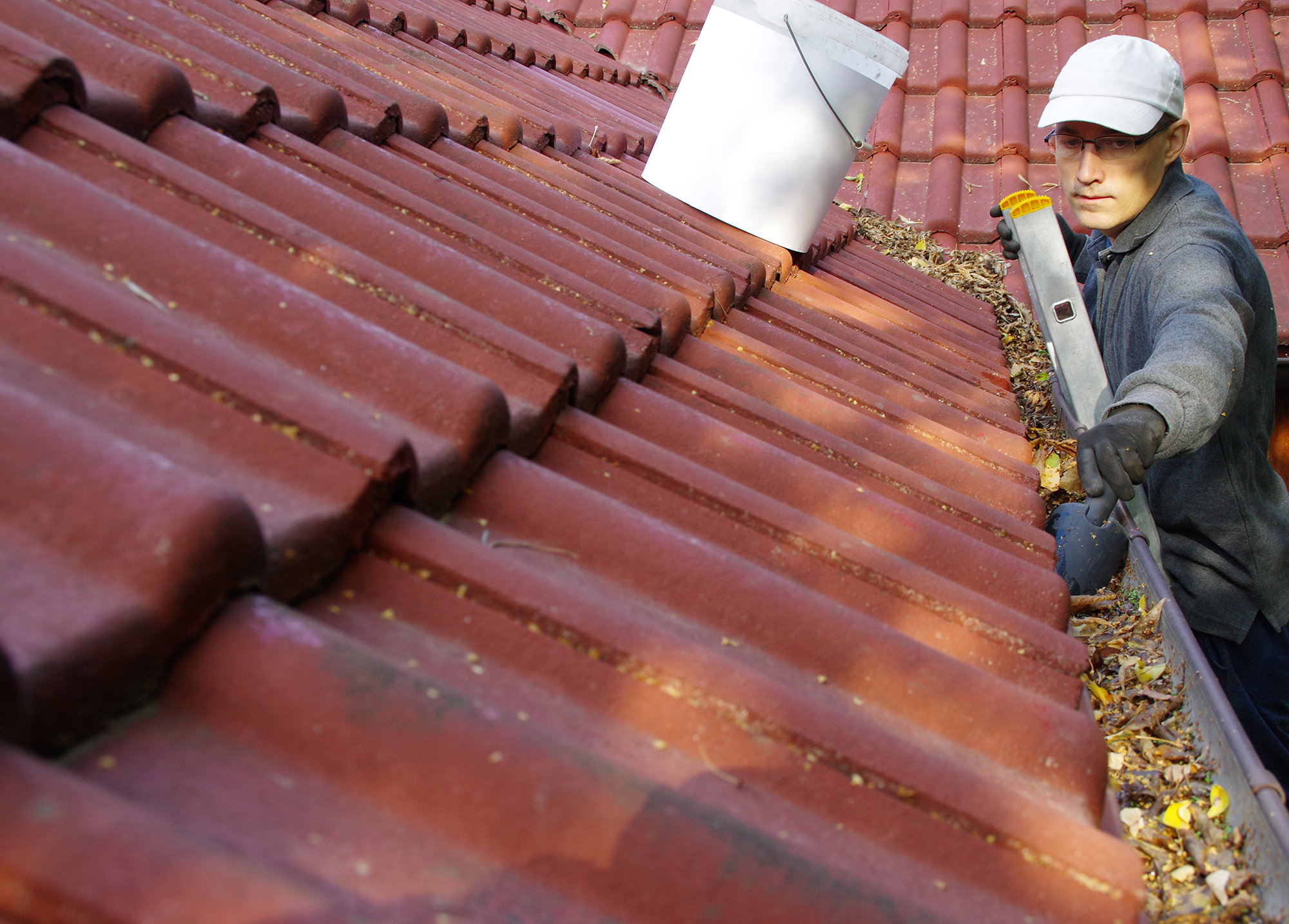 A man on a ladder with a scoop that he is using to get leaves out of clogged gutters and dump the debris into a bucket to dispose of.