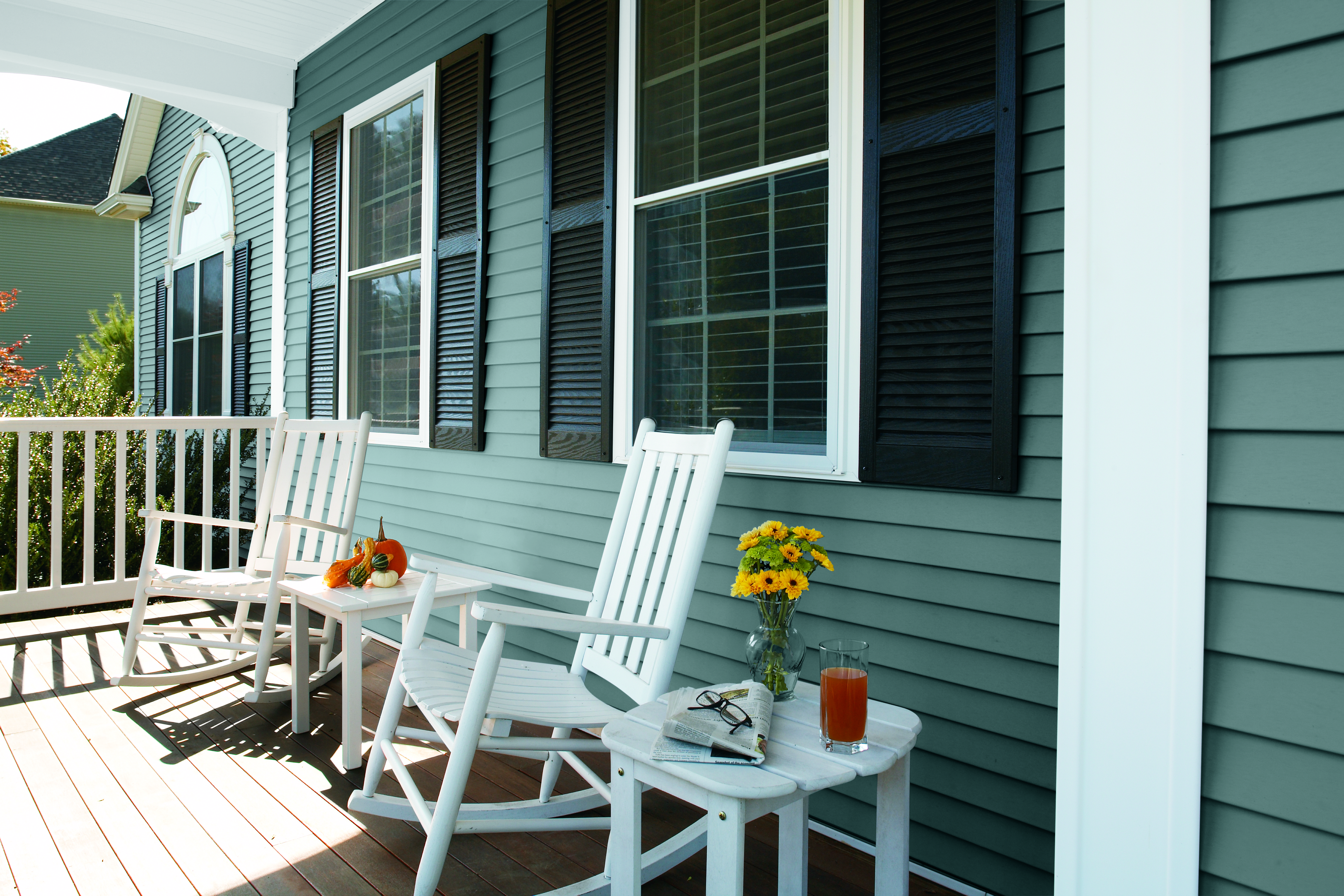 The front porch of a house with deep blue vinyl siding, large windows with black shutters, and white rocking chairs for decoration.