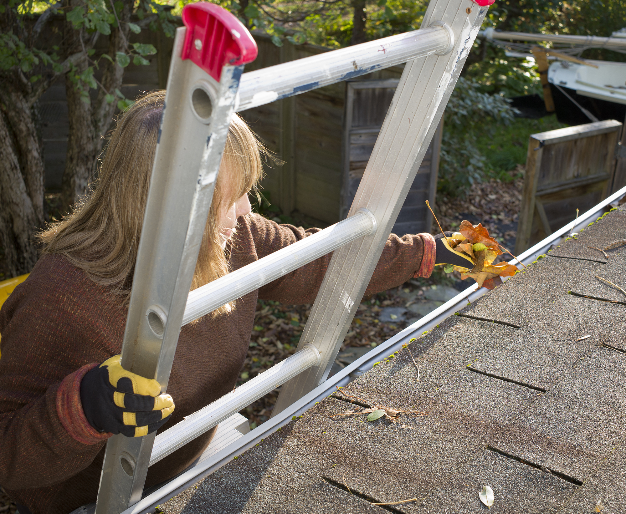 A woman on a ladder with gloves on as she cleans leaves and debris out from the gutters of a home.